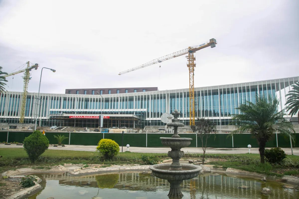 A large building with windows and white beams, two yellow construction cranes, trees, a pond and a stone fountain in front of it. 