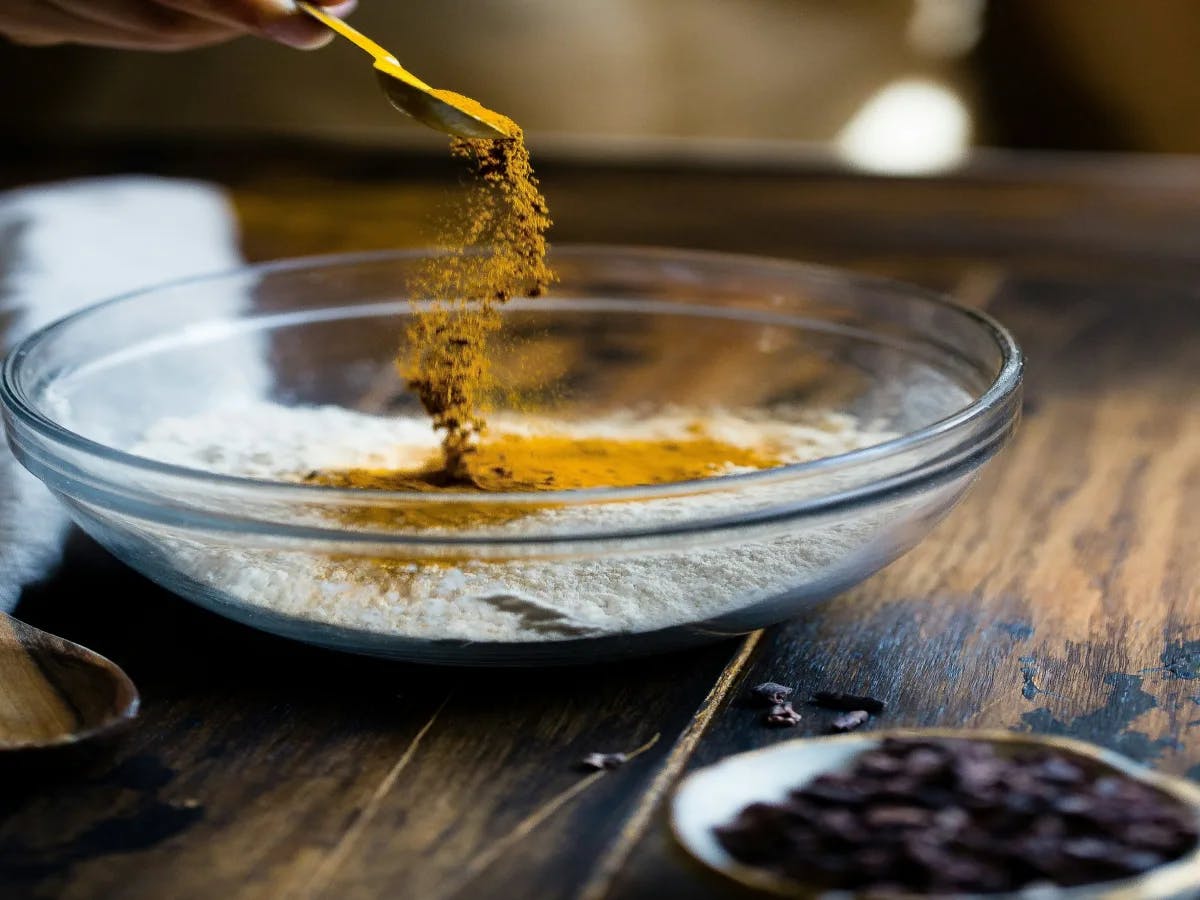 The image captures a moment of cooking, with a hand dusting yellow powder into a bowl of white powder against a kitchen backdrop.