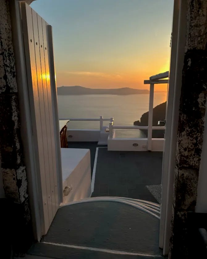 An open window looking out onto a stone patio, water and sunset with mountains in the distance