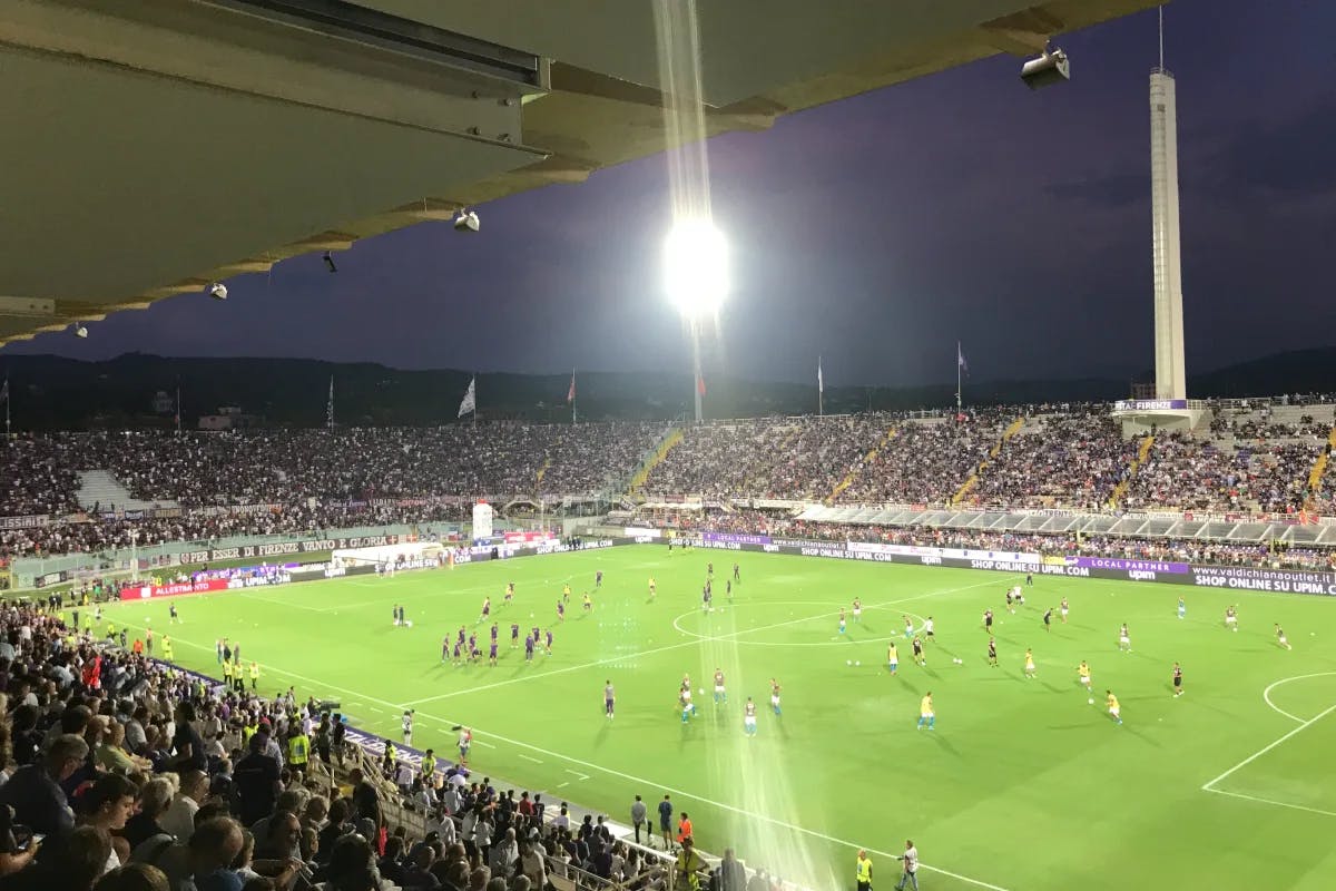 A stadium filled with people and the players on the field, playing soccer at nighttime.