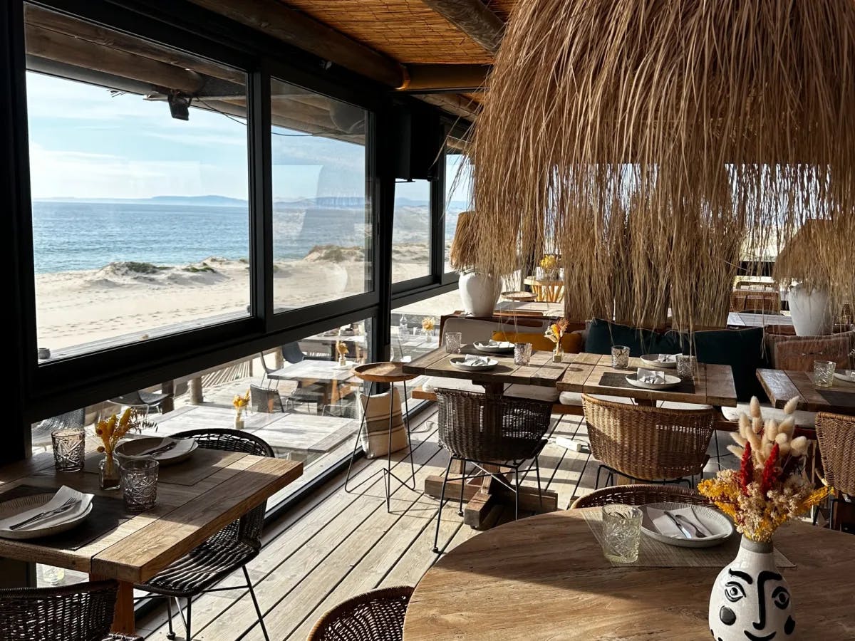 A restaurant with straw umbrellas, tables, chairs, looking out to a view of the sea and beach. 