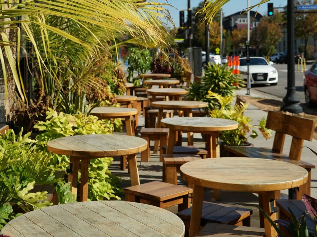 A view of wooden round tables and chairs near palm trees and bushes. 