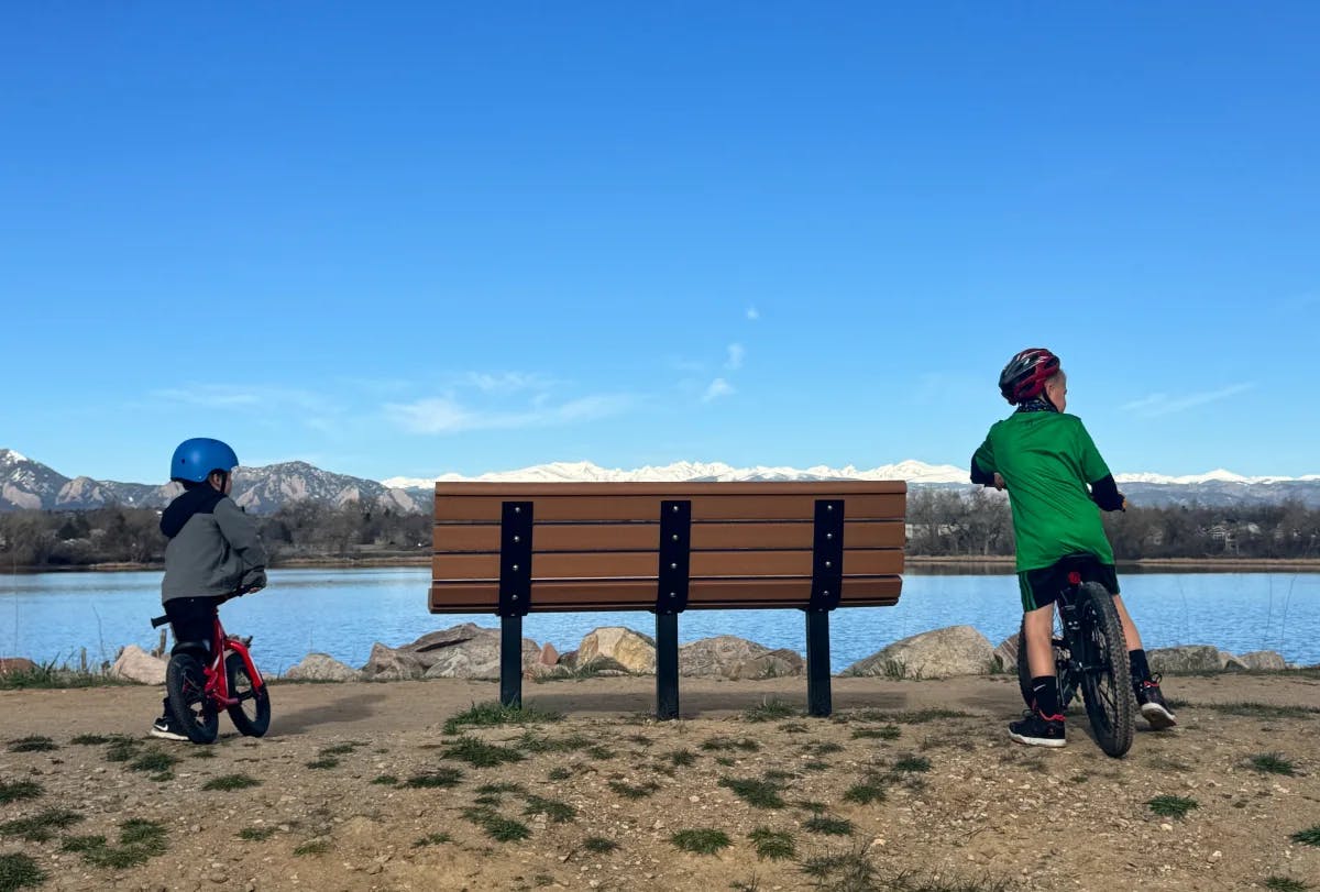 A view of two children on bicycles at the lake with the mountains of Colorado in the distance. 