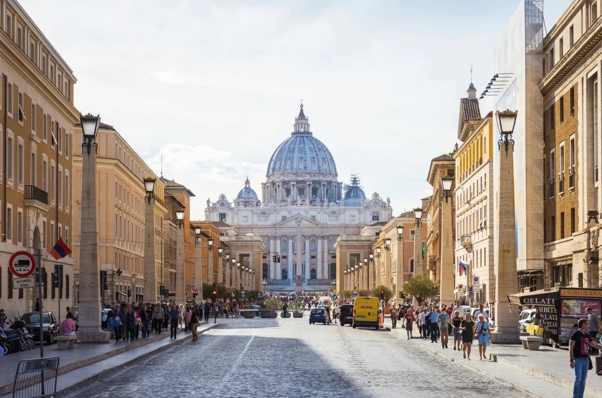 A view of a street lined with elegant buildings leading up to an impressive domed Pantheon in Rome.