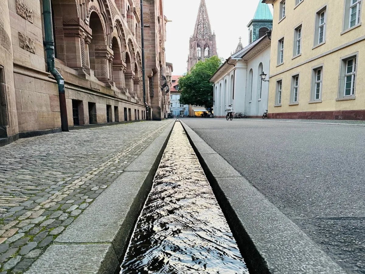 A cobblestone path leads to a distant spire bordered by historic facades and a quaint water channel evoking the charm of old European towns.