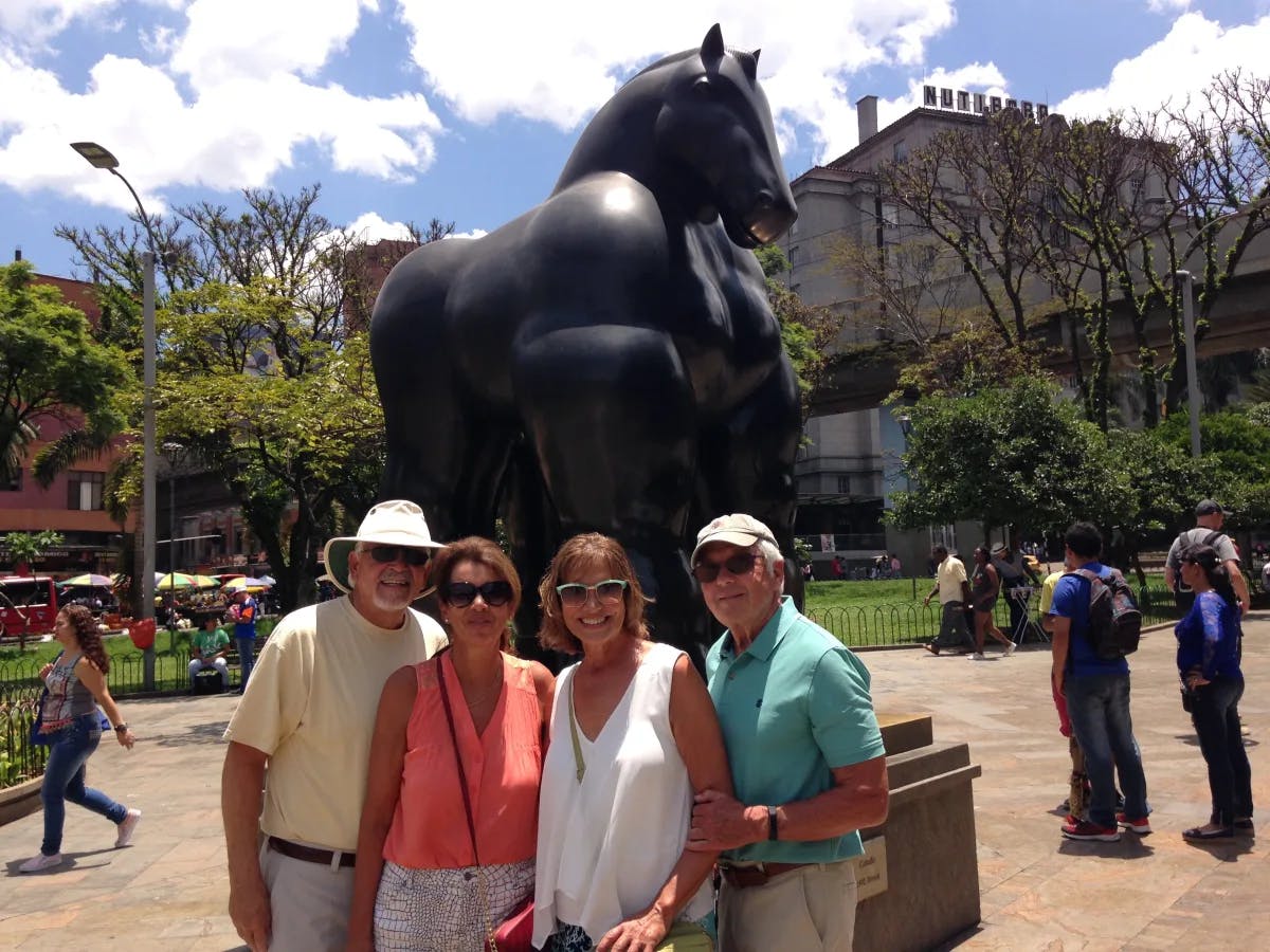 A lively quartet poses before a grand black horse statue, set against a backdrop of trees and passersby.