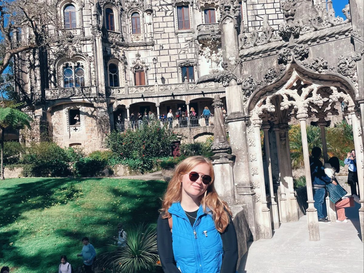 A person stands before an ornate, gothic-style building with intricate architectural details, surrounded by visitors under a clear blue sky.