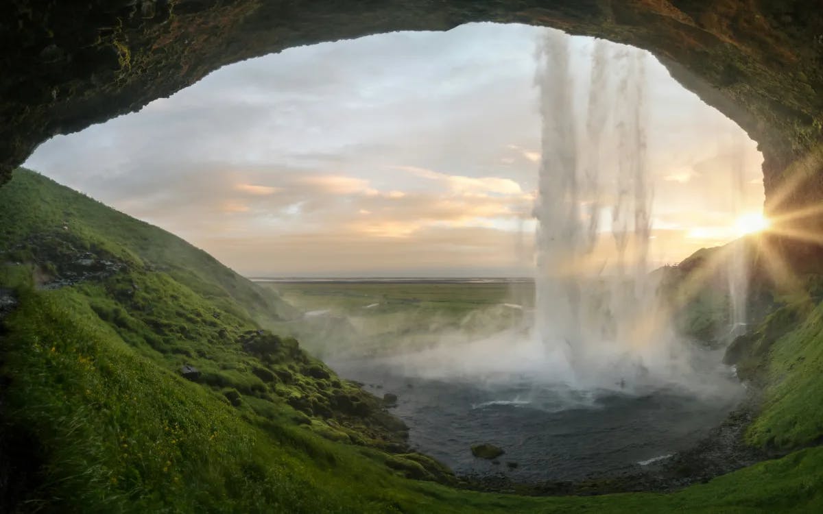 Seljalandsfoss waterfall from the inside, a cave with green hills behind the waterfall looking at the sunset.