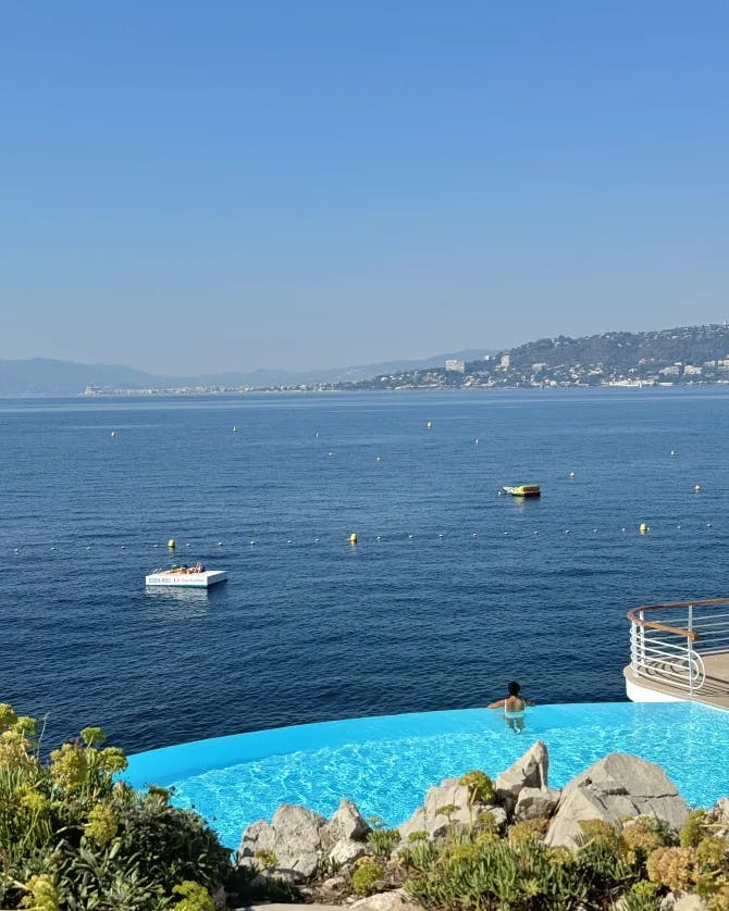 view of an infinity pool, and the sea, with buoys, and a mountainous coastline in the background.