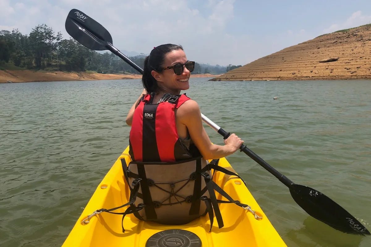 A woman in a red life-vest paddling a yellow kayak on a river with dirt banks on either side.