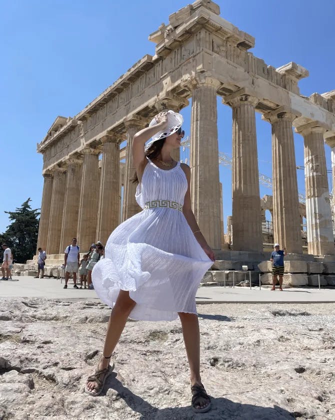 Alayna in a white dress and hat standing in front of the Acropolis on a sunny day