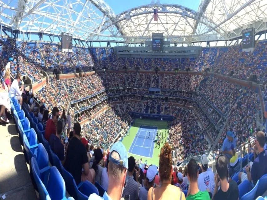 The image presents a panoramic view of a bustling tennis stadium bathed in sunlight during a match.