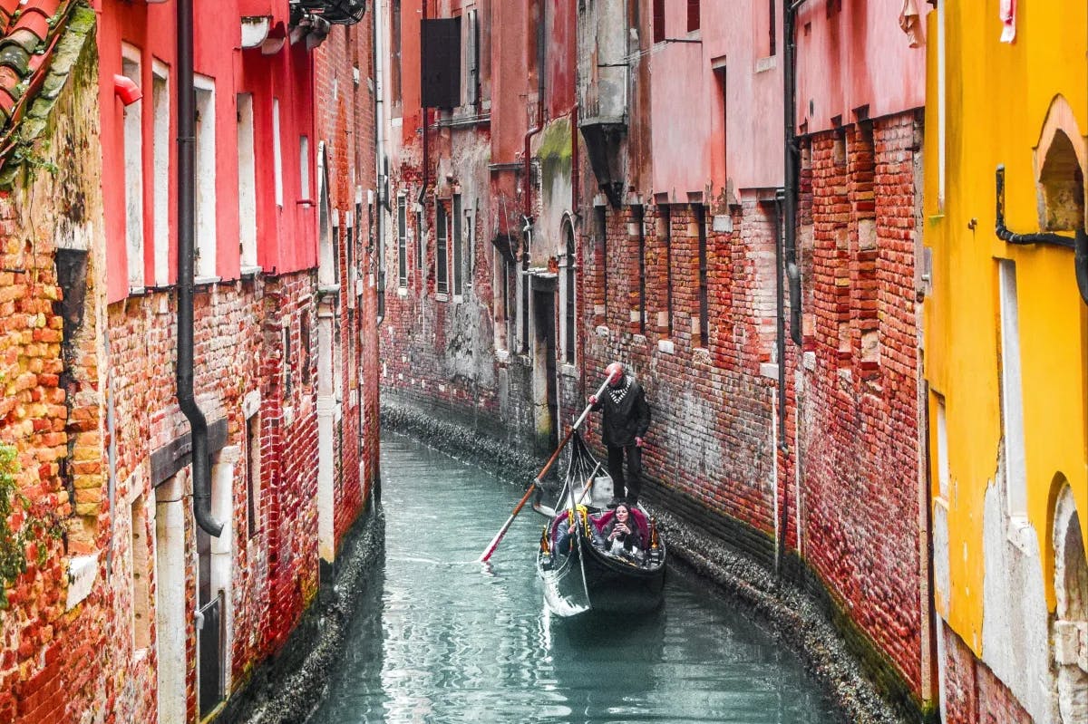 A man riding a gondola on the Venice Canal between pink and yellow brick buildings.