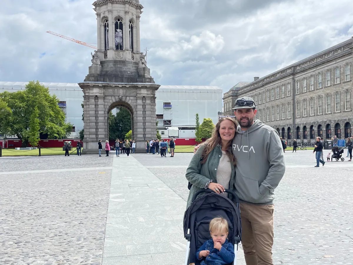 The image shows three individuals, two adults and a child in a stroller, posing for a photo with a historic tower and cloudy sky in the background.