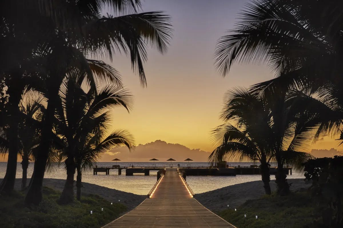 At sunset with clouds in the distance, Four Seasons Resort Nevis' private dock sits at the end of a wooden pier with palm trees surrounding the entrance