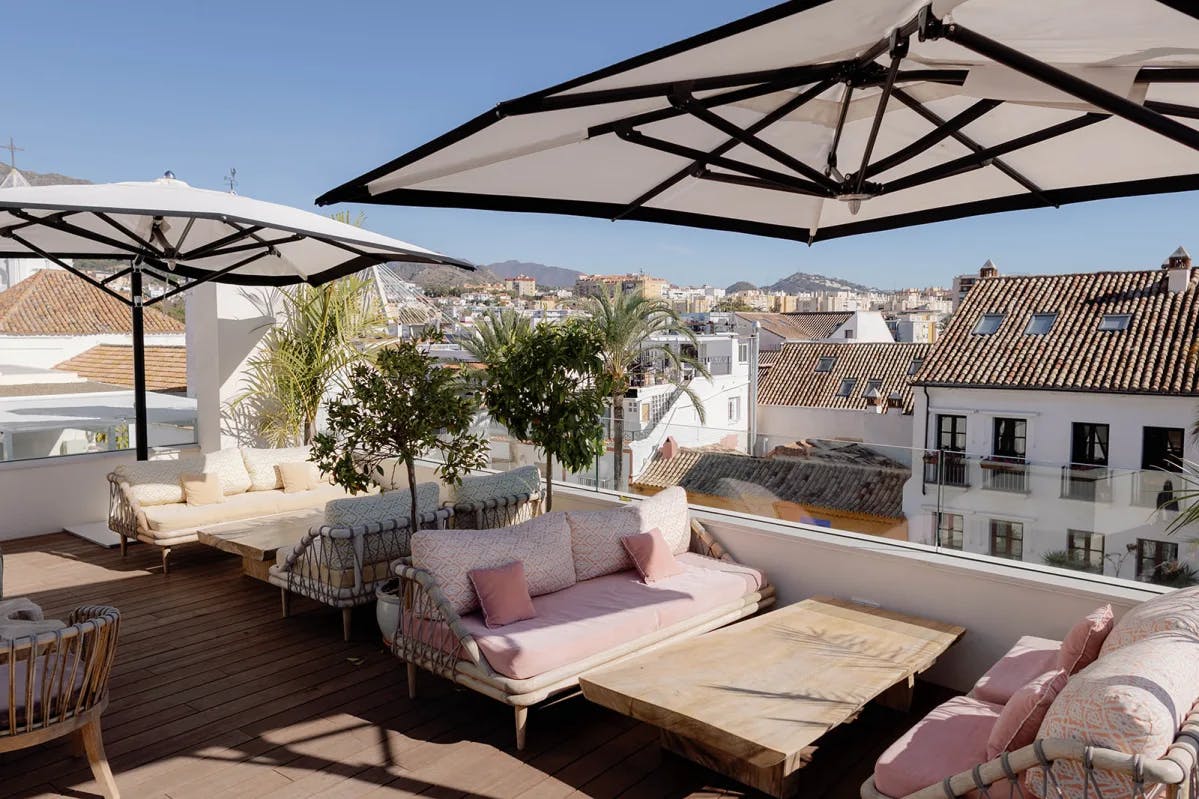 A rooftop bar with lounge seating and large white and black umbrellas, overlooking the quaint old town of Marbella and surrounding hills.