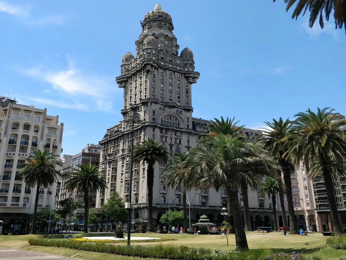 The image features a tall, ornate clock tower surrounded by a park with palm trees, set against a clear blue sky.