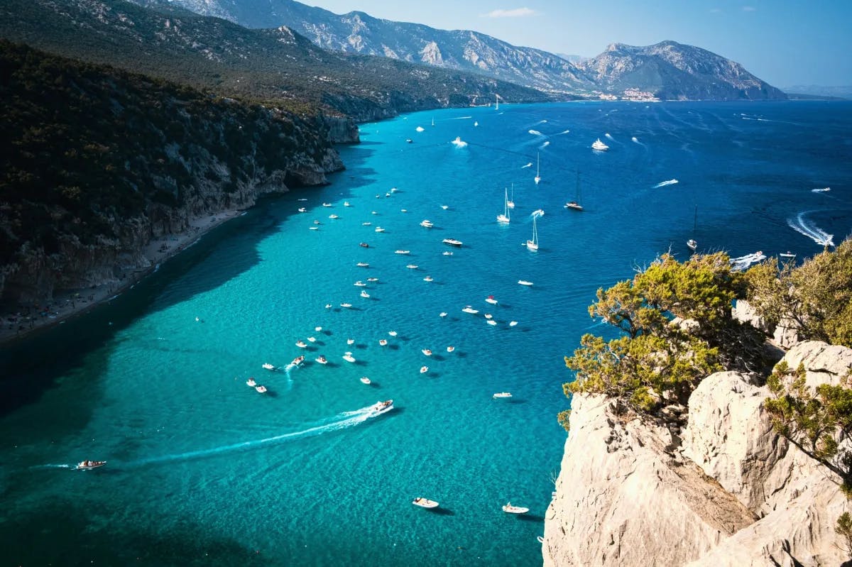 An aerial view of the turquoise sea, full of white boats and surrounded by mountains.