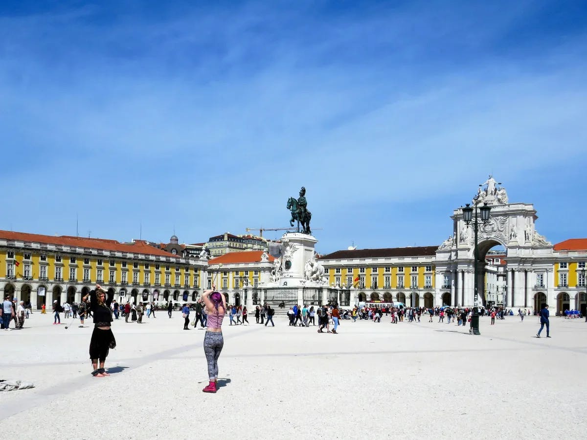 A vibrant public square bustling with people, encircled by elegant classical architecture and with a central statue.