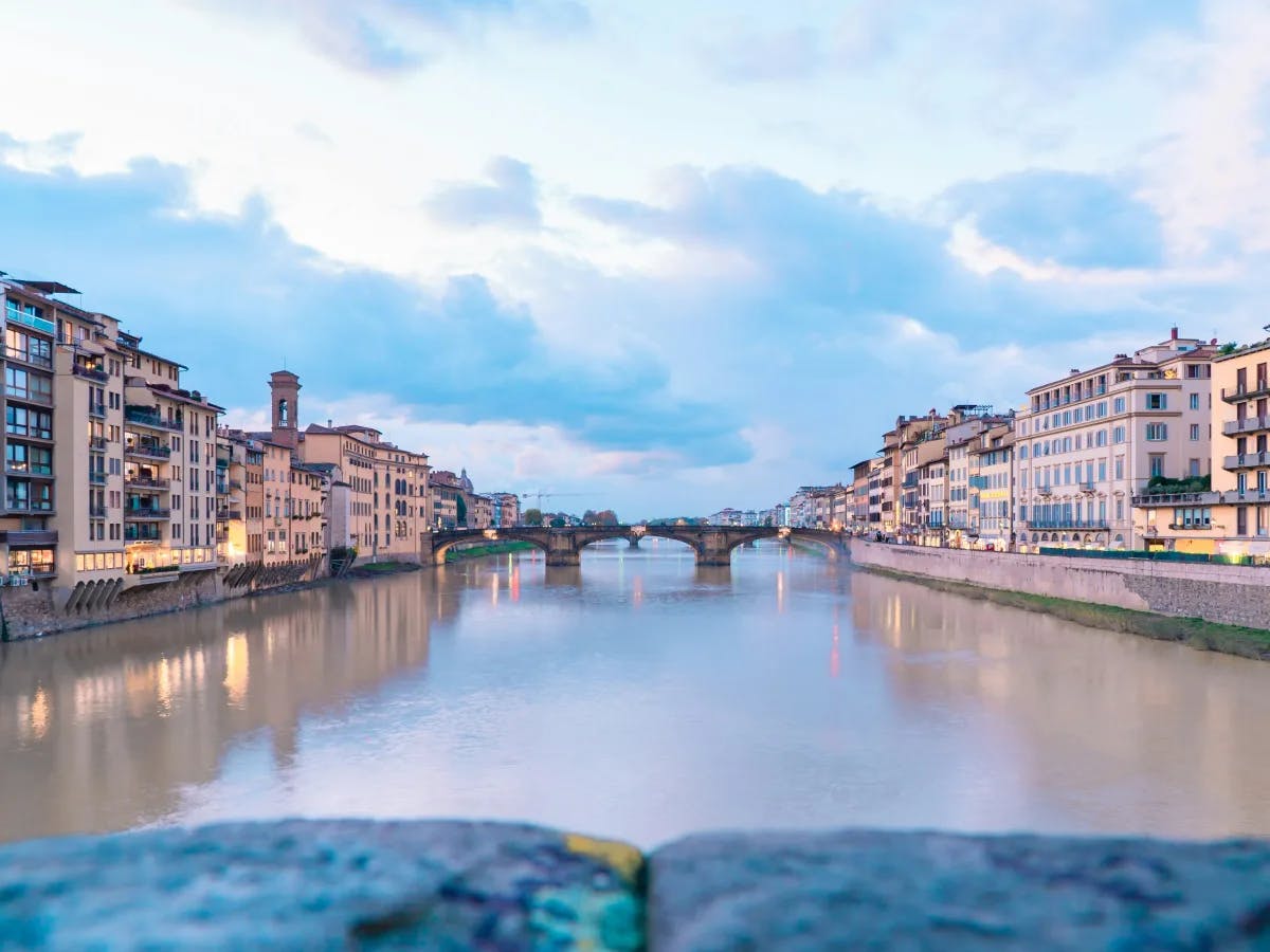 A serene view of a cityscape at dusk or dawn, with a calm river flowing through the center, flanked by buildings on both sides and a distant bridge