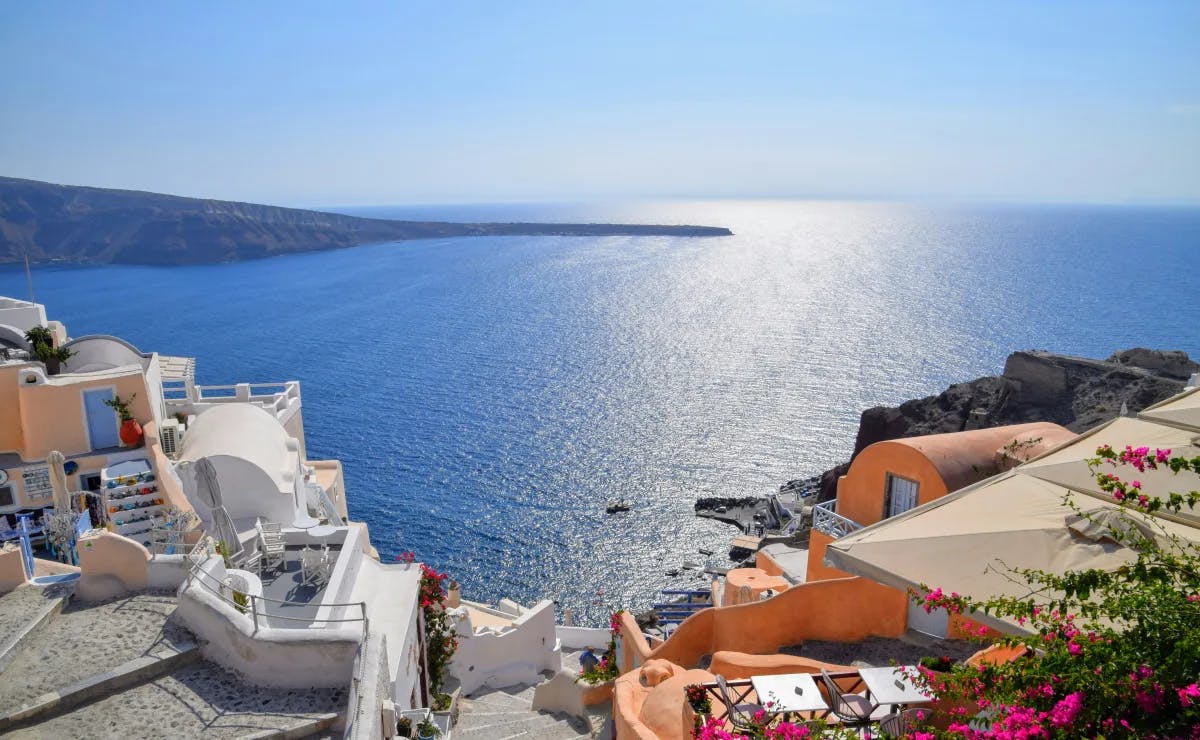 A stunning view of orange and white Greek buildings overlooking the shimmering blue sea. There is a strip of land in the distance towards the left. 