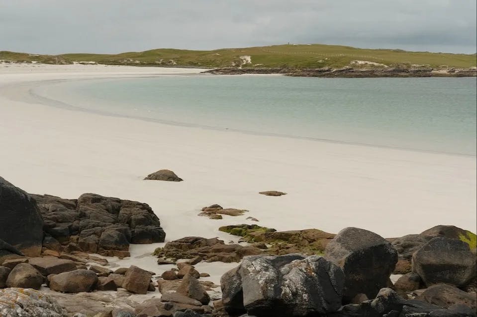 A calm ocean laps on the white sand shores of of Dog's Bay Beach on a grey day.  Green hills in the distance.