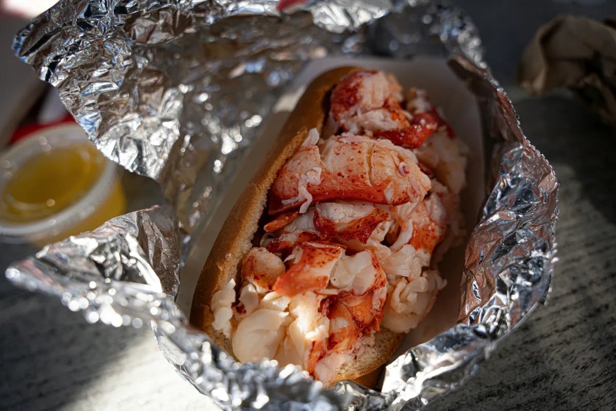 The image highlights a lobster roll, ready to be enjoyed, wrapped in foil with a side of butter, set on a speckled counter.