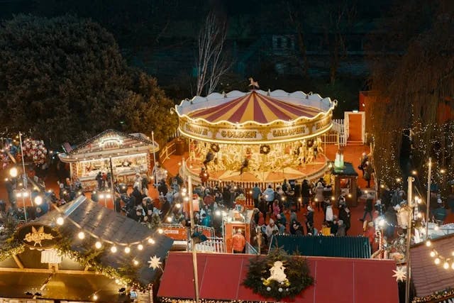 An aerial shot of a christmas fair at night with an illuminiated merry-go-round and many people walking around with string lights. 