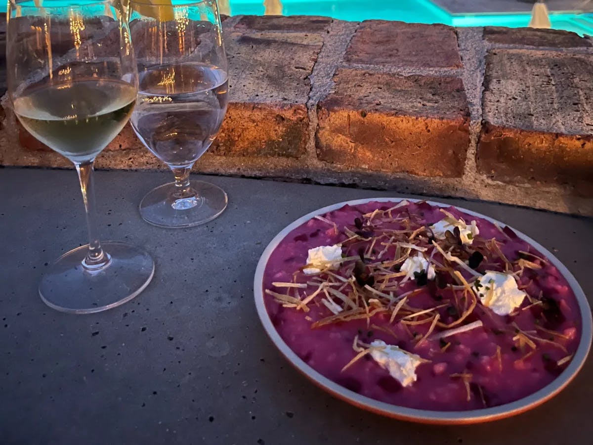 A view of purple food plated and positioned on a cement table top. There is a glass of wine and water to the left of it, and bricks in the background. 