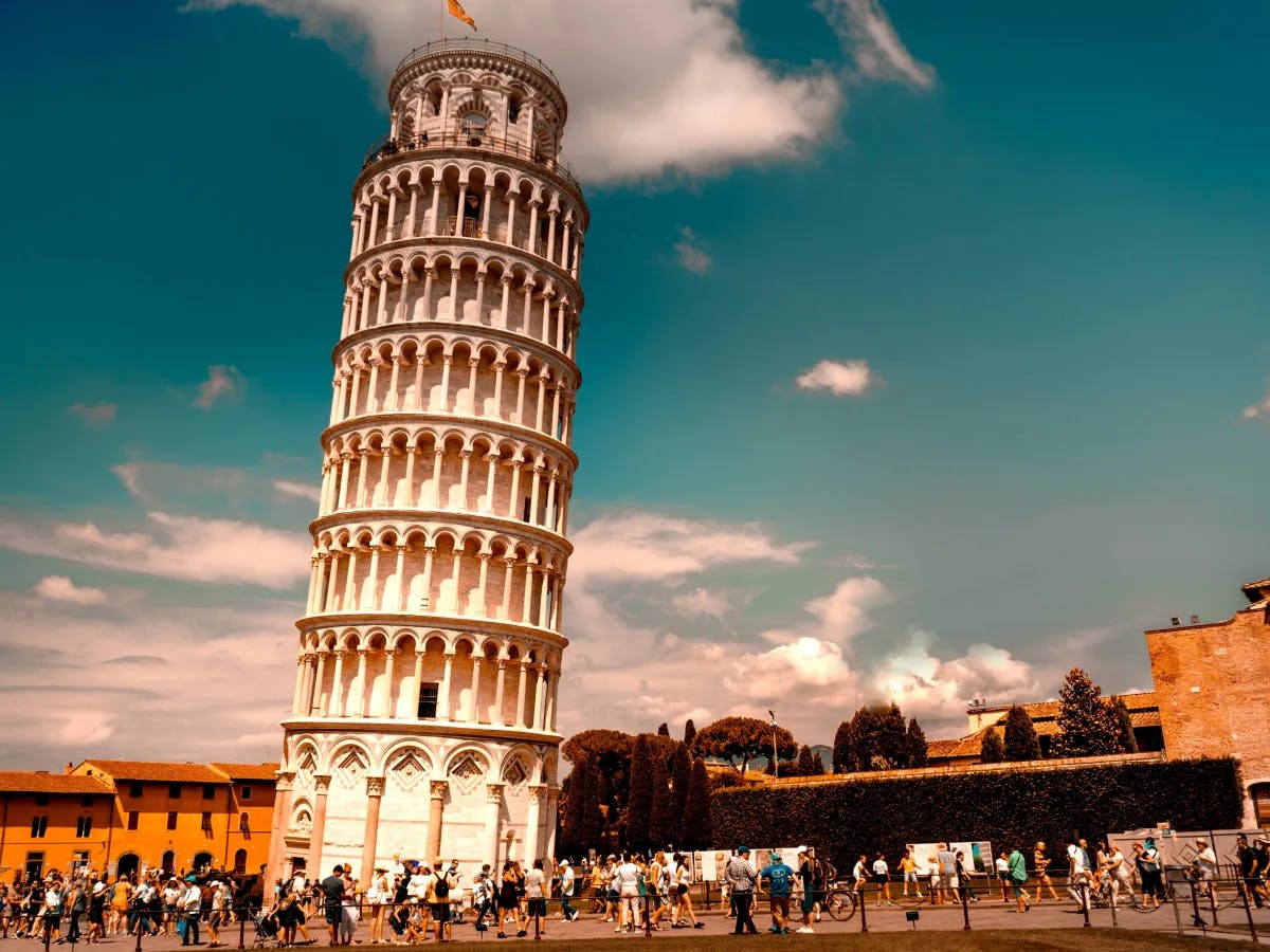 Image of the Leaning Tower of Pisa reaching into the sky on a sunny day dotted with clouds as tourists mill below.