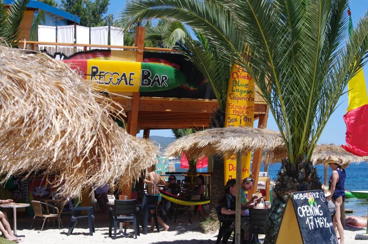 A nice view of the entrance to Reggae Beach Bar with palm trees and outdoor seating on the beach. 