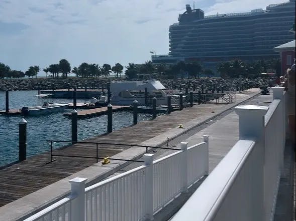 This image depicts a wooden dock with boats and a large cruise ship in the background. There is white fencing in the forefront and green trees in the distance. 