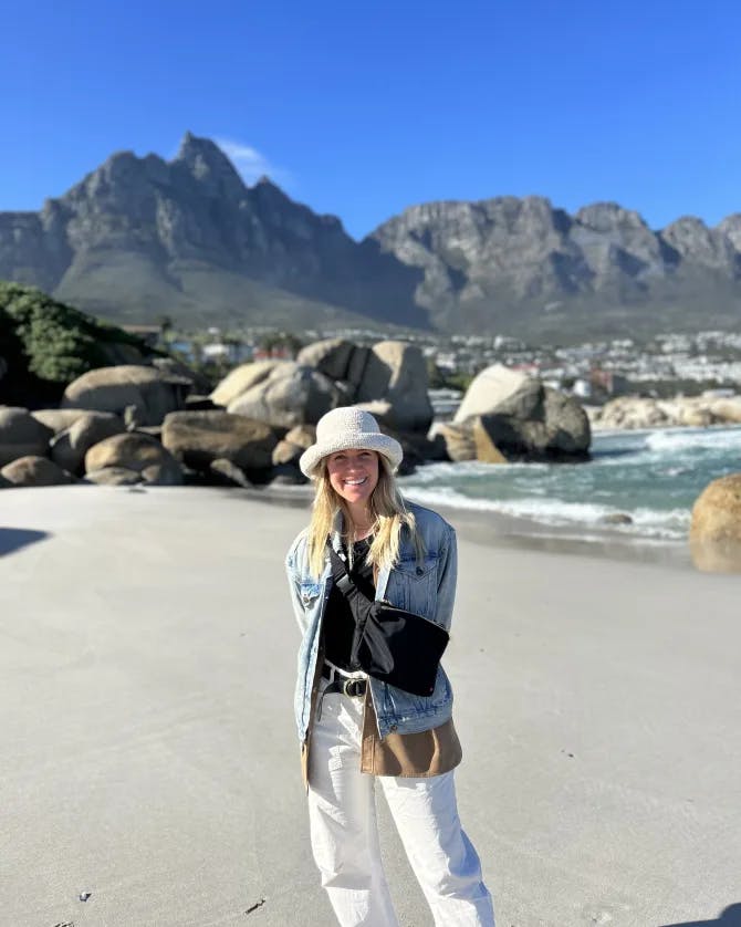 Molly in a white bucket hat standing on a beautiful empty beach with a mountain landscape visible behind her and clear skies