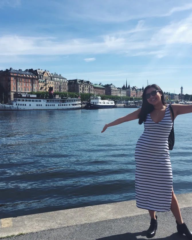 A woman in black and whit striped dress in front of a city lake with buildings behind. 