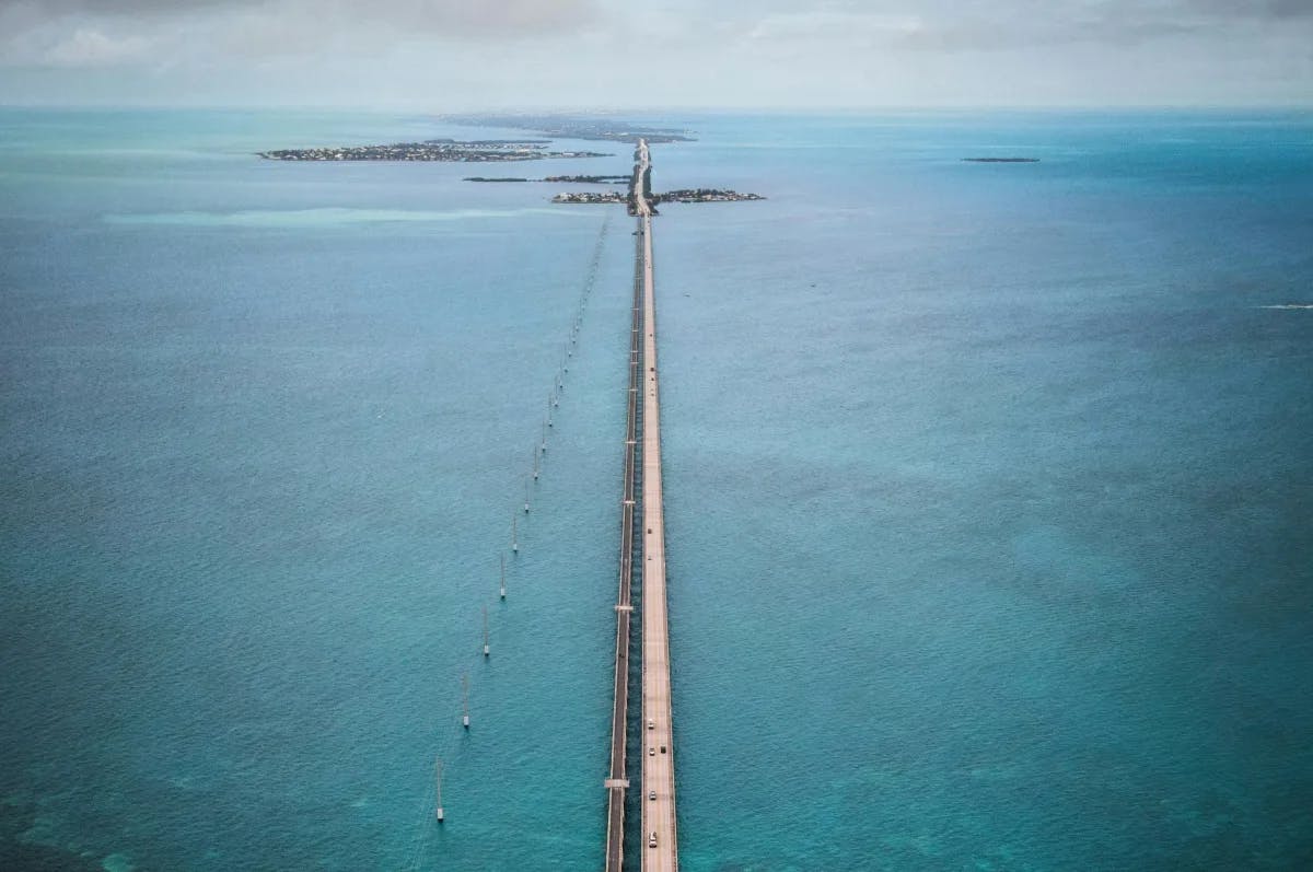 An aerial view of a long narrow bridge with cars driving surrounded by ocean