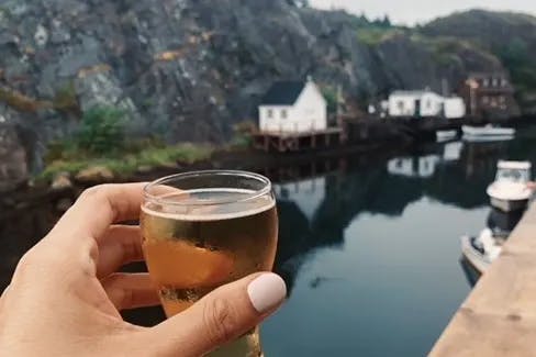 A hand holding a frosty glass overlooking a harbor and mountains in the distance on an overcast day. 