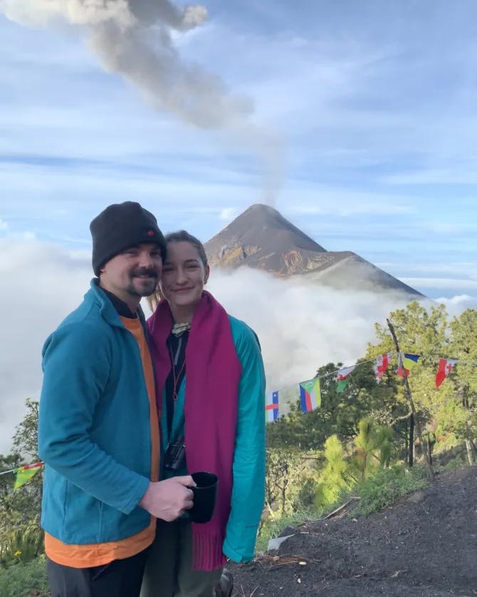 Shelby posing with her partner in front of a volcano and green bushes 