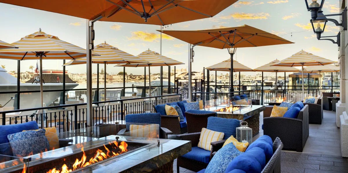 A restaurant patio at the Balboa Bay Resort with outdoor firepits, umbrellas and lounge-style seating with blue and yellow cushions.