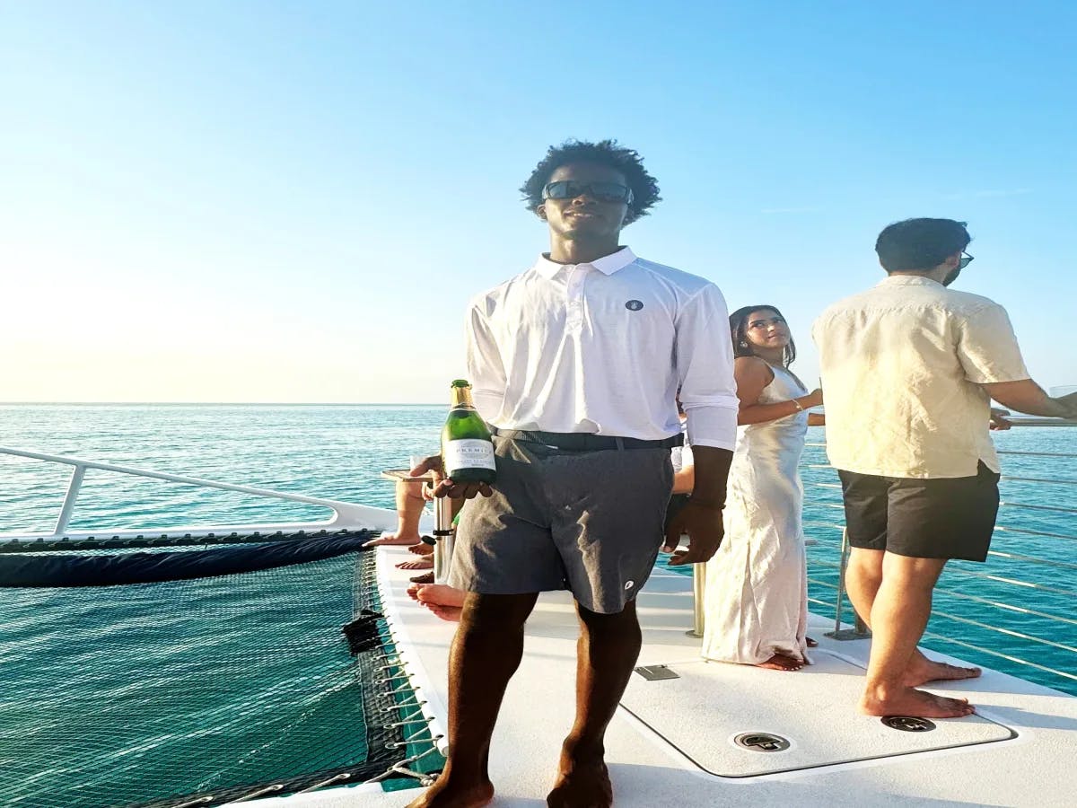 The image depicts a man holding a bottle of champagne on a boat with people admiring the ocean view behind.