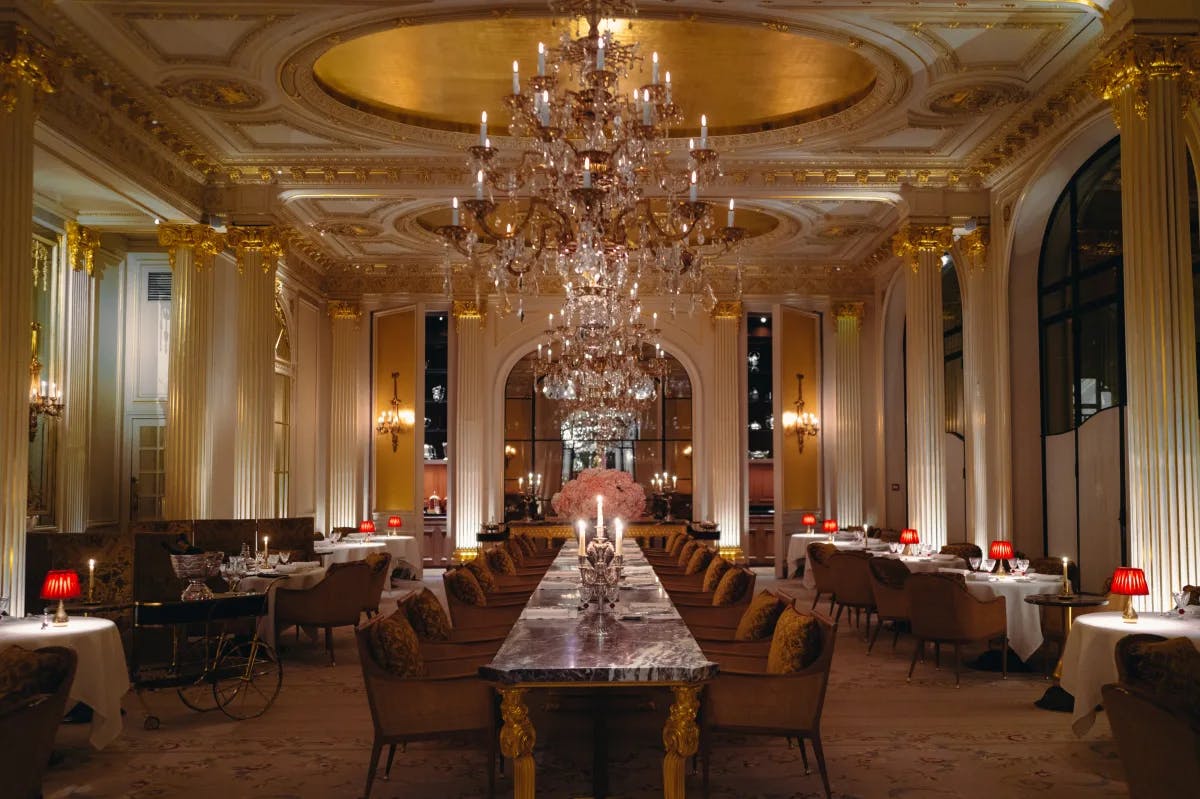 A restaurant at Plaza Athénée hotel with very elegant and plush interior featuring a long table in the middle underneath a grand chandelier. The room has dim lighting with gold fixtures.