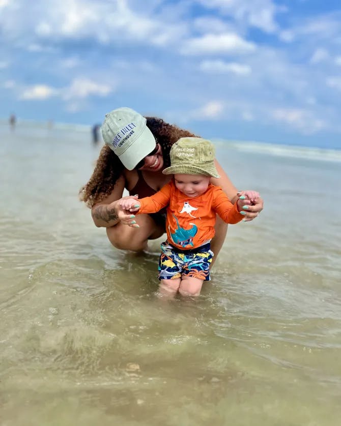 A woman posing in the ocean with a small baby in her arms and smiling.