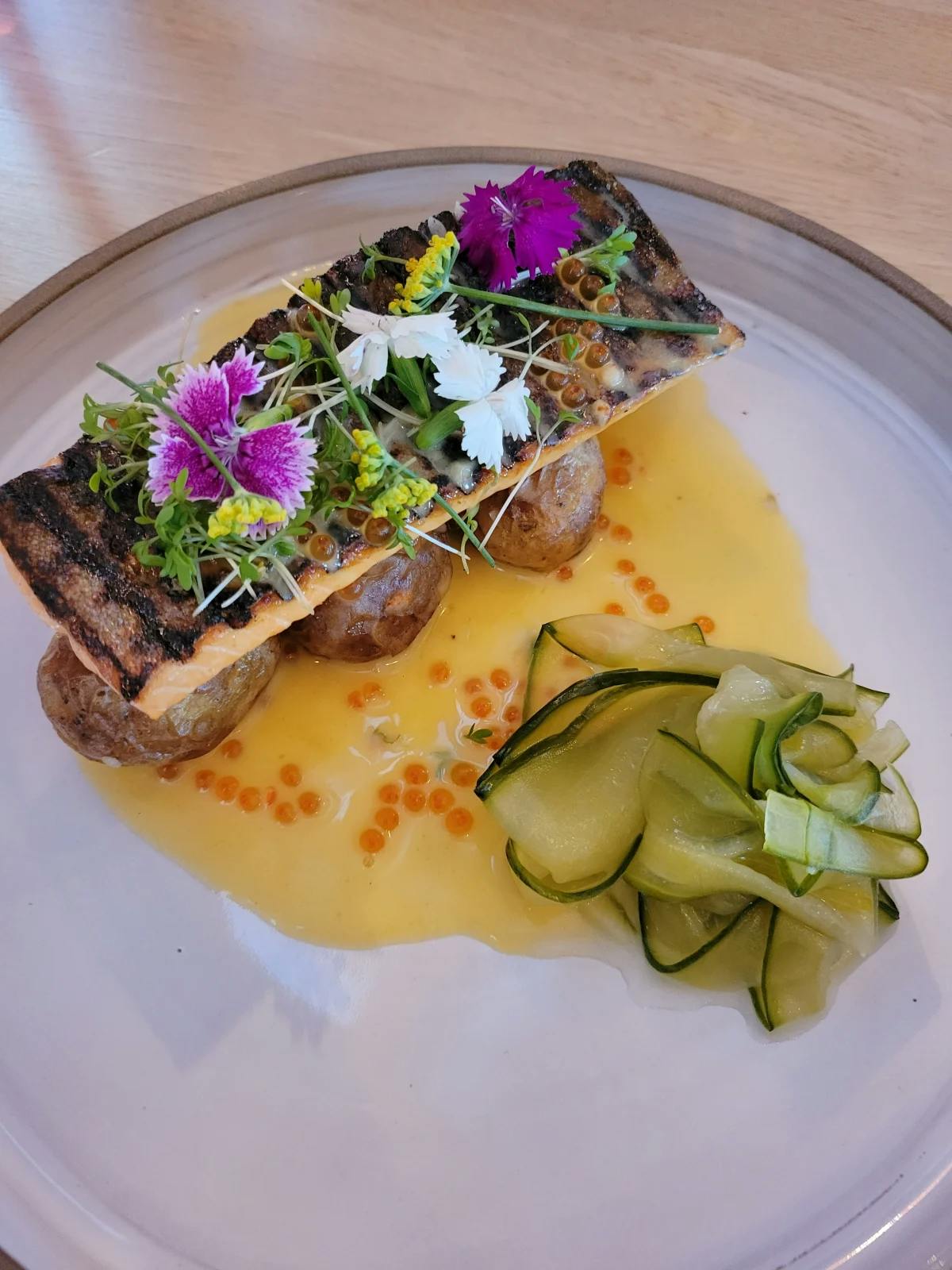 An elegant plate of grilled salmon topped with colorful flowers and resting on potatoes, with a thinly-cut cucumber salad, a yellow sauce and orange caviar.