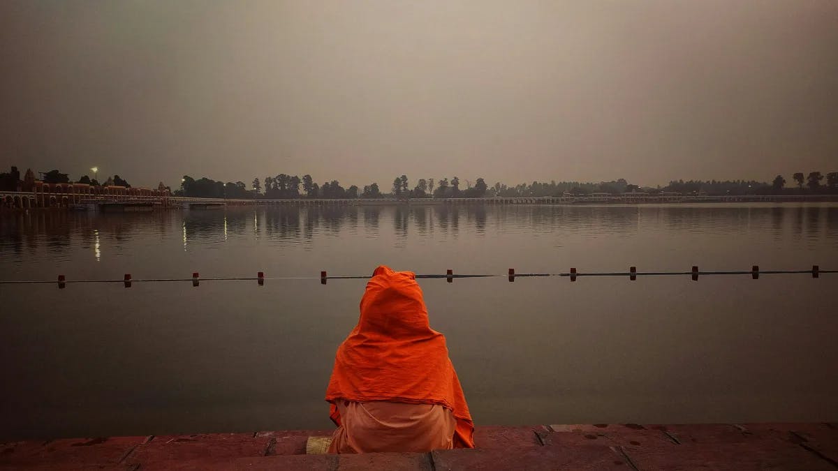 A person in orange wearing a headscarf sitting at the edge of a large lake.