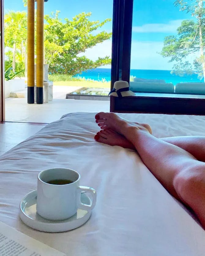 A person having tea in bed with view of the ocean outside their window.