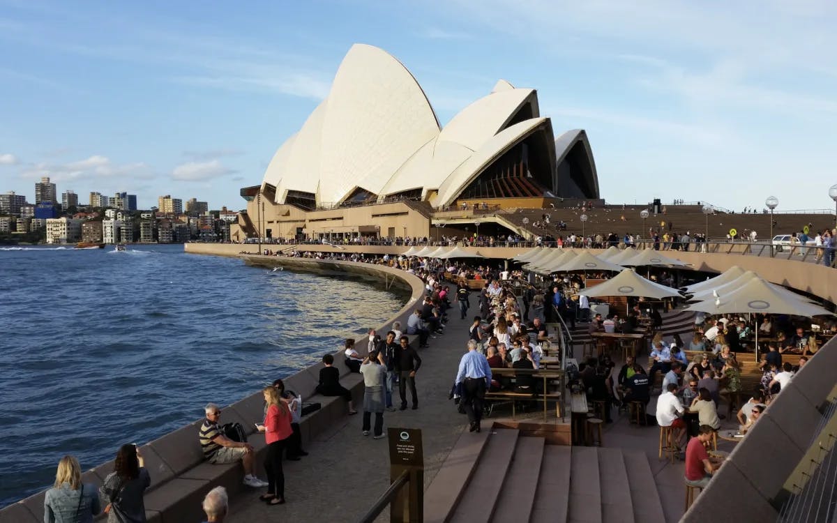 A view of people dining under umbrellas near a boardwalk in front of the Sydney Opera House near the blue water.