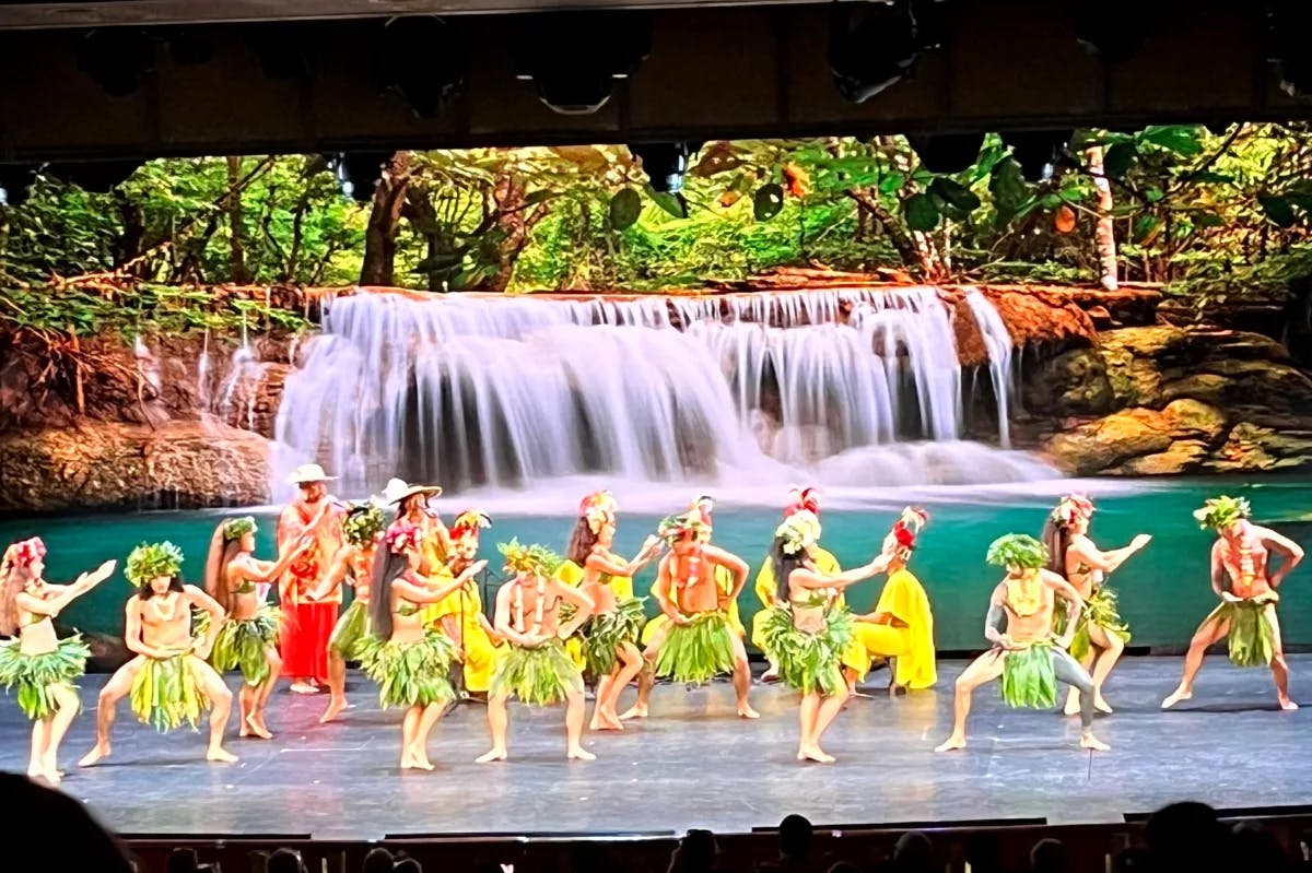 A group of cultural dances performing on a stag with an image of a waterfall behind them 