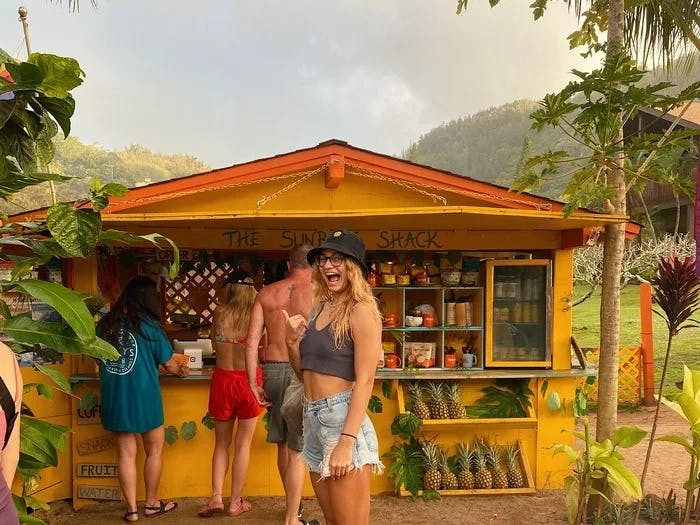 The image shows a vibrant yellow food shack named “The Sunrise Shack,” with people standing in front, in a tropical setting.