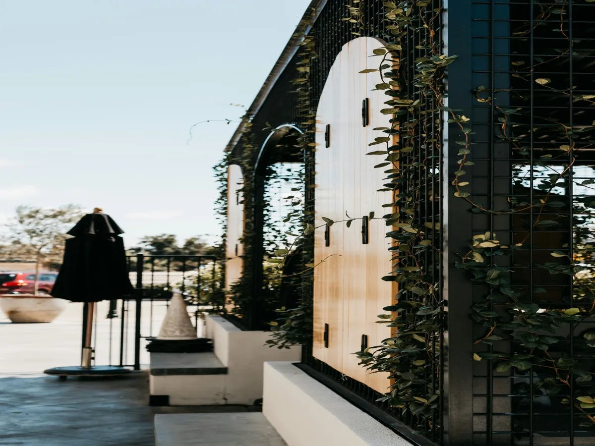 A modern building facade with arched windows covered by a black grid and climbing plants, blending architectural and natural elements.