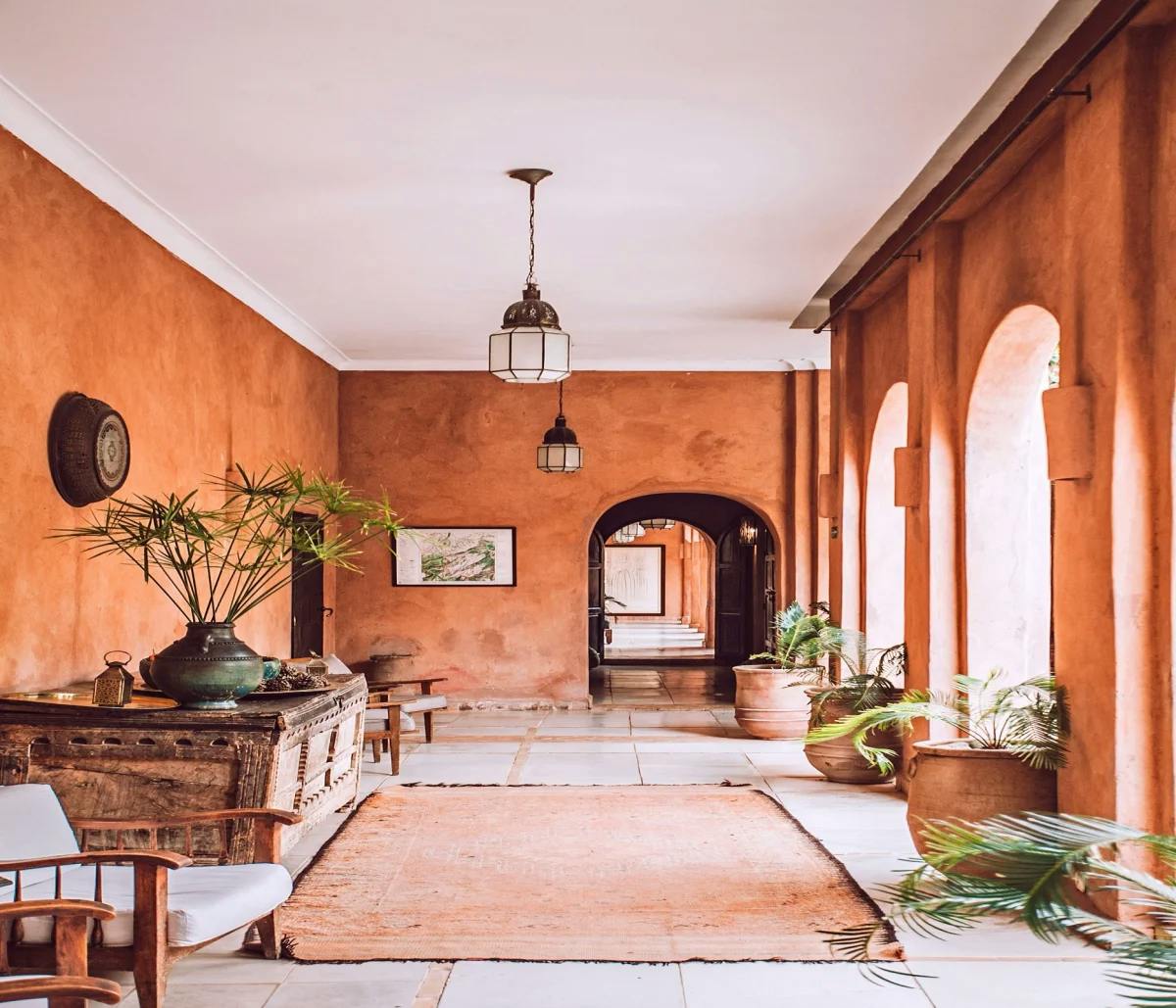 An orange walled corridor with arches, hanging chandeliers, a rug and plants near wood and wicker furniture. 