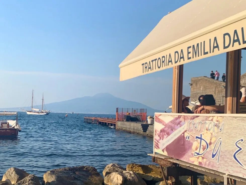 The image presents a tranquil seaside view with the sign “TRATTORIA DA EMILIA DAL 1947” and boats on the water, suggesting a charming dining spot by the sea.